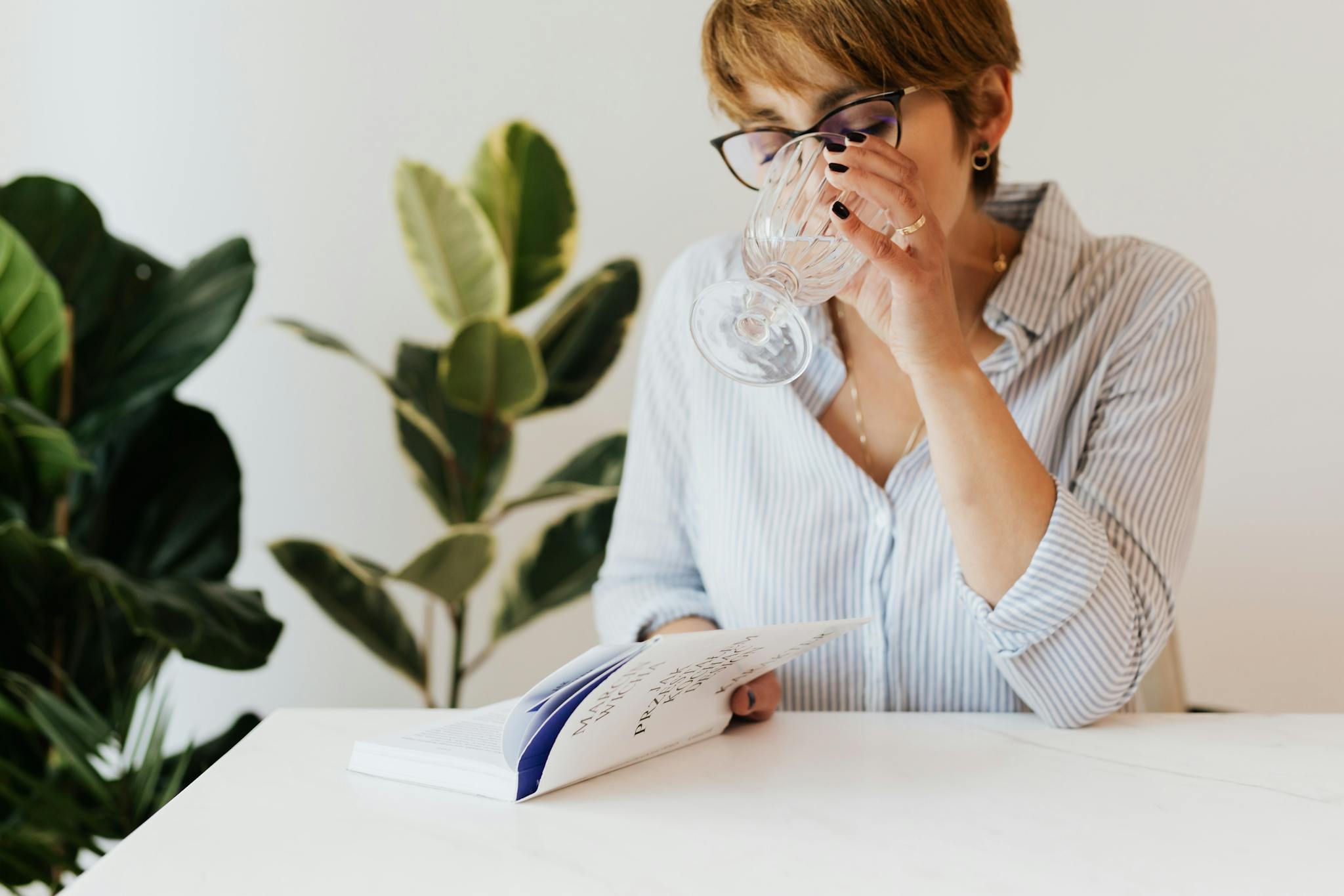 A woman enjoys a book and a refreshing drink indoors, surrounded by plants, capturing leisure and relaxation.